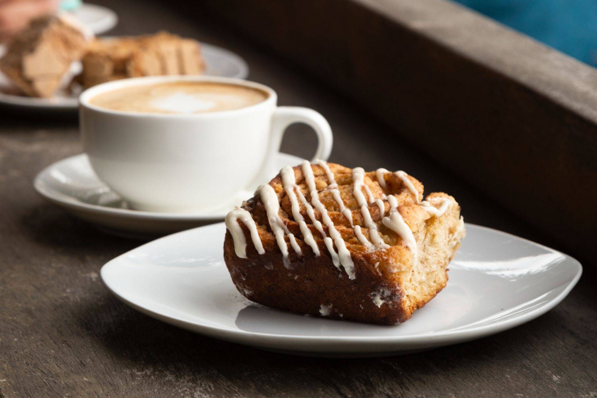 Eine weiße Tasse mit Kaffee, neben einem Stück Kuchen mit Zuckerglasur auf einem weißen Teller.